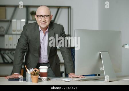 Portrait d'un cadre caucasien mûr souriant en costume formel debout à la table avec ordinateur dans le bureau moderne Banque D'Images