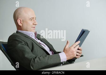 Content décontracté homme d'affaires chauve dans la veste assis dans la chaise de bureau et d'utiliser une tablette tout en travaillant en ligne au bureau Banque D'Images