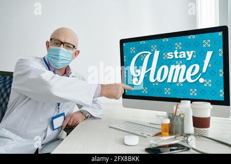 Portrait de médecin chauve mature stricte en masque et blanc manteau assis au bureau et pointant vers l'écran d'ordinateur pendant vous demandant de rester à la maison Banque D'Images