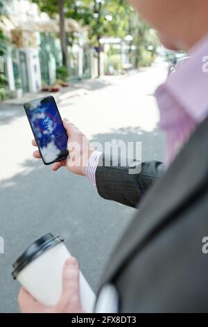 Vue sur l'épaule d'un homme méconnaissable en veste buvant un café et vérifier les prévisions météorologiques au téléphone tout en marchant le long de la rue Banque D'Images