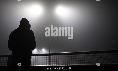 Un mystérieux, solitaire figure à capuche, de retour à l'appareil photo. Debout sur un pont, sur une autoroute en hiver brumeux la nuit. Banque D'Images