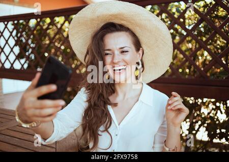 happy modern femme de ménage d'âge moyen en chemise blanche avec chapeau ayant la web cam réunion sur un smartphone dans la terrasse. Banque D'Images