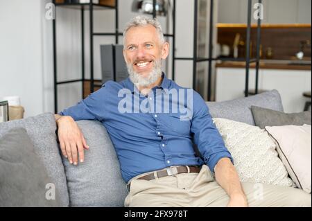 Beau gris mûr homme plein de personnalité assis sur le canapé dans une posture détendue, regarde loin et sourit. Style haut de la main à l'œil bleu style senior élégant et décontracté Banque D'Images