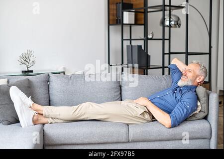 Un petit garçon hippster aux cheveux gris serein repose sur le canapé dans le salon confortable, un homme mûr allongé sur le canapé, se détendre à la maison, un homme âgé souriant reflétant, rêvant de jour sur le canapé Banque D'Images