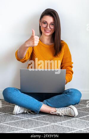 Une jeune femme utilise un ordinateur portable assis sur un tapis blanc et en montrant le pouce vers le haut Banque D'Images