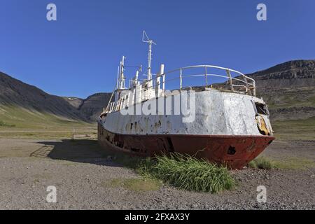 Coque blanche érodée d'un vieux bateau de pêche abandonné reposant sur un sol graveleux dans une vallée montagneuse des Vestfjords en Islande, ciel d'été clair Banque D'Images