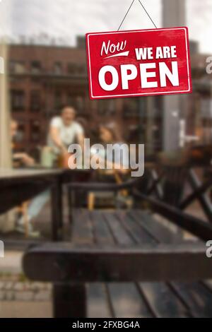 Maintenant - nous sommes signe ouvert devant des gens flous à l'extérieur d'un café appréciant le dîner, les boissons et le café. Les bars et les restaurants ouvrent après Coronaviru Banque D'Images