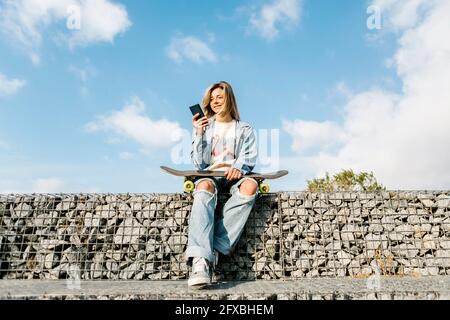 Femme souriante utilisant un téléphone portable assis sur un mur de pierre Banque D'Images