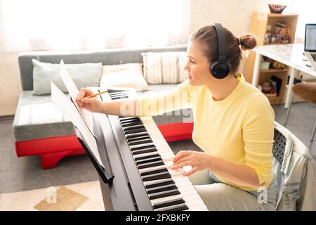 Femme adulte de taille moyenne portant un casque d'écoute écrivant tout en apprenant le piano à la maison Banque D'Images