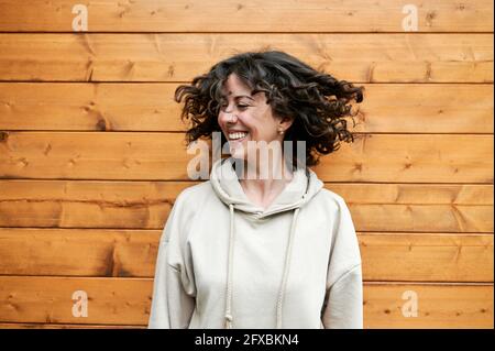 Femme souriante se jetant les cheveux tout en se tenant devant un mur en bois Banque D'Images