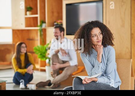 Femme d'affaires moyenne adulte attentionnés qui regarde loin tout en étant assise avec des collègues en arrière-plan au bureau Banque D'Images