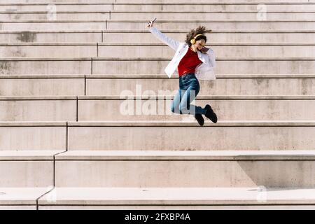 Femme ecstatique avec casque et smartphone sautant dans l'escalier pendant la journée ensoleillée Banque D'Images