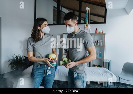 Thérapeute physique féminine avec un objet crâne debout collègue mâle tenant des poids devant la table de massage en pratique pendant une pandémie Banque D'Images