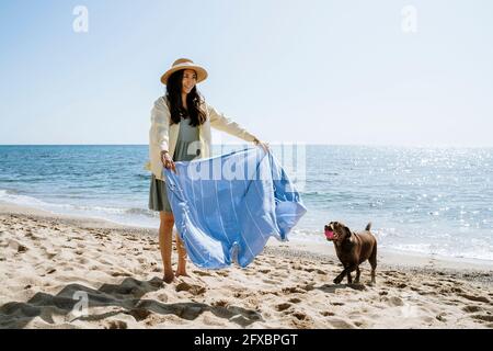 Femme souriante tenant un chiffon près d'un chien sur le sable à la plage Banque D'Images