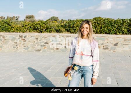 Femme souriante avec planche à roulettes debout dans le parc pendant la journée ensoleillée Banque D'Images