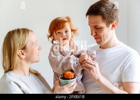 Une mère souriante avec un cupcake regardant la fille portée par son mari dans la cuisine Banque D'Images