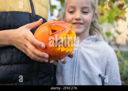 Enfant tenant et ouvert au-dessus d'une citrouille sculptée avec un visage ghoulish, Jack-o-lanterne, dans le fond garçon assis à la table et décorant la citrouille Banque D'Images