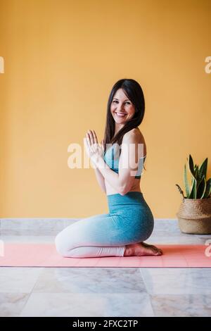 Femme gaie aux mains claspétées pratiquant le yoga sur un tapis d'exercice à la maison Banque D'Images