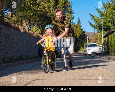 Un père souriant aide sa fille à faire du vélo sur la route Banque D'Images