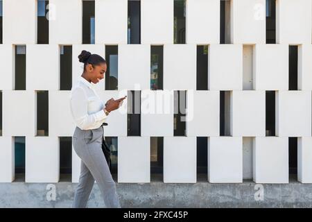 Femme d'affaires souriante utilisant un téléphone portable tout en marchant près du mur Banque D'Images