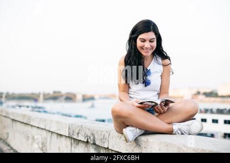 Femme adulte de taille moyenne souriant en lisant un livre assis sur un mur de soutènement Banque D'Images