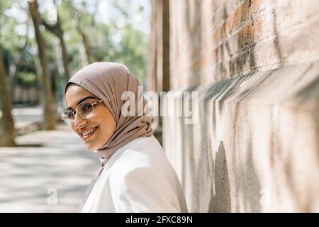 Femme gaie portant des lunettes de vue debout près du mur pendant la journée ensoleillée Banque D'Images