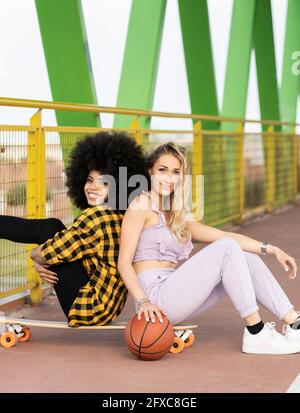 Des amies de sexe féminin s'asseyant dos à dos sur une planche à roulettes au pont Banque D'Images