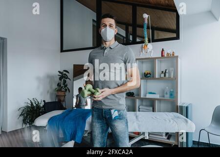 Un physiothérapeute masculin portant un masque facial protecteur tenant des poids dans la pratique Banque D'Images