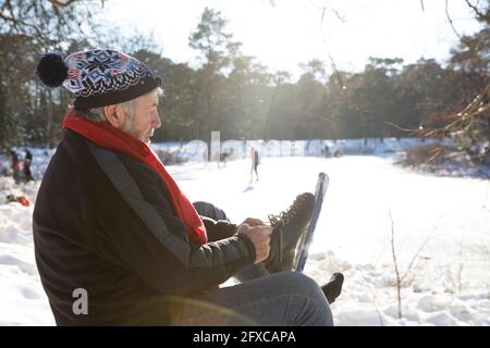 Homme âgé portant un patin à glace tout en étant assis sur la neige pendant l'hiver Banque D'Images