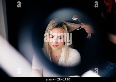 Coiffeuse féminine utilisant un lisseur sur les cheveux du magnifique modèle vu à travers l'anneau lumineux Banque D'Images