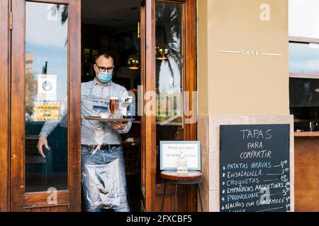 Un garçon portant un masque facial tenant des boissons dans un plateau pour les clients à l'extérieur d'un bar Banque D'Images