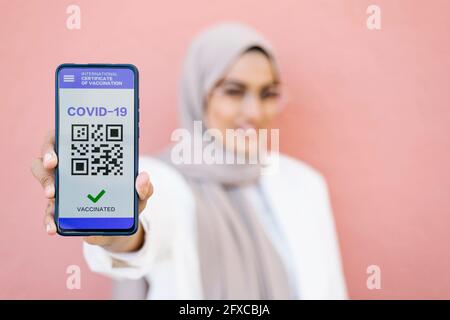 Jeune femme montrant un certificat de vaccination COVID-19 sur smartphone devant le mur Banque D'Images