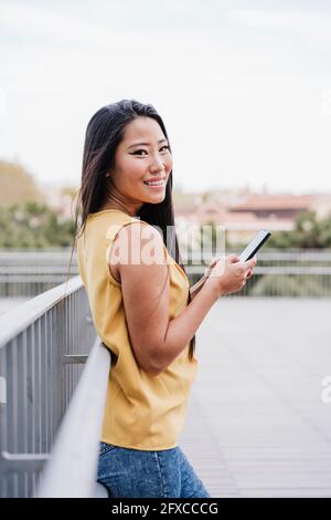 Femme souriante tenant un smartphone tout en s'inclinant sur une rambarde à une passerelle surélevée Banque D'Images