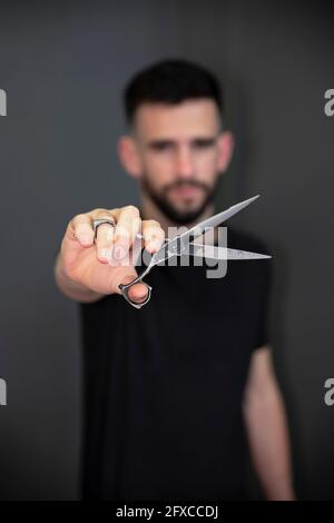 Jeune homme barber montrant le ciseau en face du mur noir au salon Banque D'Images