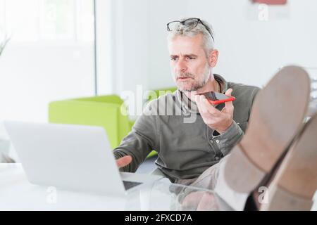 Homme entrepreneur sérieux travaillant sur ordinateur portable tout en envoyant la messagerie vocale par téléphone mobile au bureau Banque D'Images