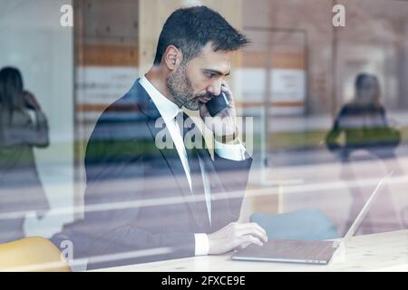 Homme d'affaires sérieux utilisant un ordinateur portable tout en parlant au téléphone au bureau Banque D'Images