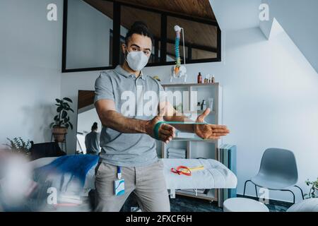Un physiothérapeute masculin portant un masque de protection tout en étirant la bande de résistance en pratique Banque D'Images