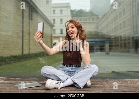 Femme en train de gesturer tout en prenant le selfie à travers le téléphone mobile devant le mur de verre Banque D'Images