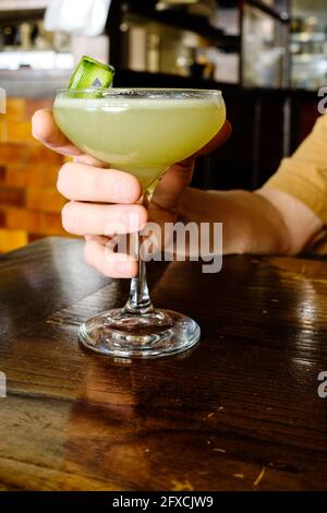 homme tenant un gimlet de concombre dans un verre à tige assis à une table en bois Banque D'Images