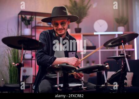 Jeune homme inspiré dans des lunettes de soleil et un chapeau tendance jouant à la batterie dans le studio sombre. Musicien talentueux créant un nouveau rythme à l'aide d'instruments électroniques. Banque D'Images