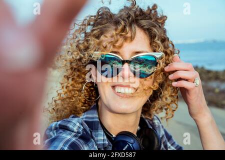 Gros plan sur les cheveux bouclés femme en lunettes de soleil faisant selfie photo sur l'appareil photo. Une jolie jeune femme avec casque prend le selfie tout en marchant près de la mer. Extérieur Banque D'Images