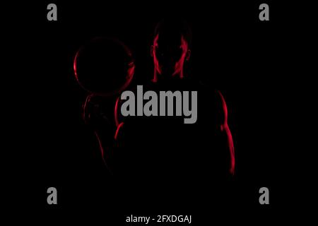 Joueur de basket-ball tournant une balle sur fond noir. Silhouette afro-américaine rouge avec éclairage latéral. Banque D'Images