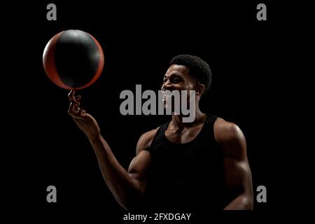 Joueur de basket-ball tournant une balle sur fond noir. Bonne silhouette d'homme afro-américain souriant. Banque D'Images