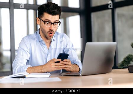 Excité surpris de jeunes caucasiens courtier, gérant ou indépendant portant des lunettes, assis à son bureau, utilisant un smartphone, choqué par les nouvelles ou le message, regardant l'écran dans l'étonnement Banque D'Images