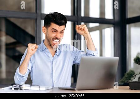 Homme d'affaires, indépendant ou gestionnaire, joyeux et réussi, assis à la table au bureau, hurle et se réjouit de la réussite, de la victoire ou du bon profit, regarde un ordinateur portable et des gestes avec ses mains Banque D'Images