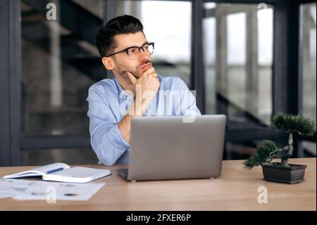 Pensive et réussie jeune chef d'entreprise, directeur ou courtier caucasien dans des vêtements et des lunettes formels, assis à un ordinateur portable dans le bureau, regardant sur le côté, reposant sa tête sur sa main Banque D'Images