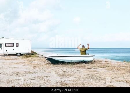 Homme assis en bateau sur la plage près de sa camionnette de camping-car blanche garée au bord de la mer. Saison touristique sur la mer méditerranée. Sicile. Mer Ionienne. Banque D'Images