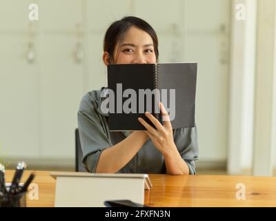 Portrait en demi-longueur d'une jeune étudiante amicale cachant son visage derrière un livre assis sur le lieu de travail Banque D'Images