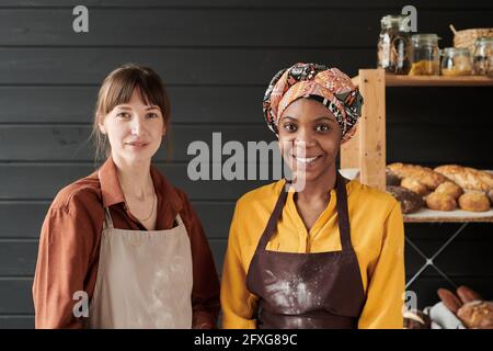 Portrait de deux jeunes boulangers en tabliers souriant à l'appareil photo tout en cuisinant dans la cuisine Banque D'Images