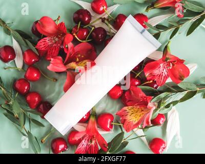 Soins de la peau à plat avec tube de crème avec cerises et fleurs dans l'eau sur fond vert naturel Banque D'Images